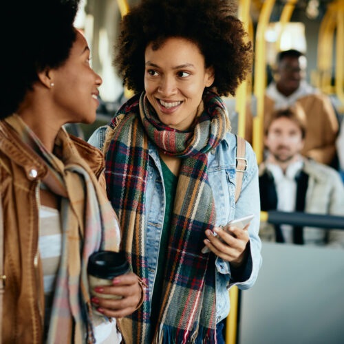 Happy black woman talking to her female friend while using mobile phone and traveling by bus.